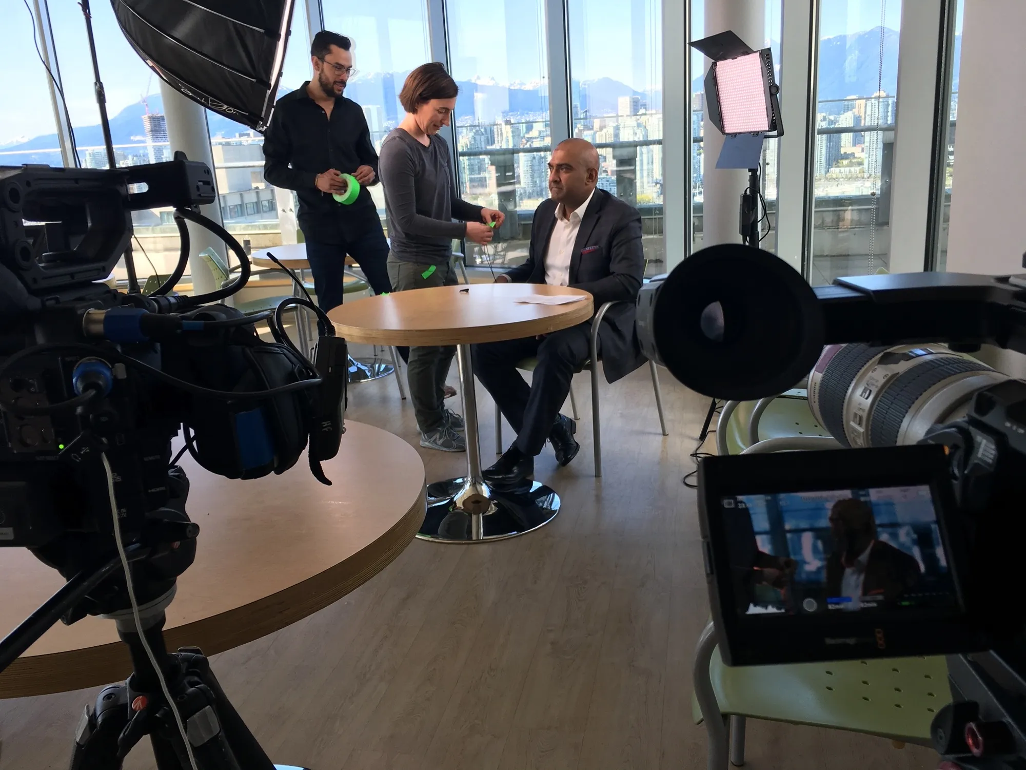On set with a Vancouver skyline backdrop: the crew mics up Microsoft’s Raj Cheema at a round table before his interview, snow-capped mountains through the glass.