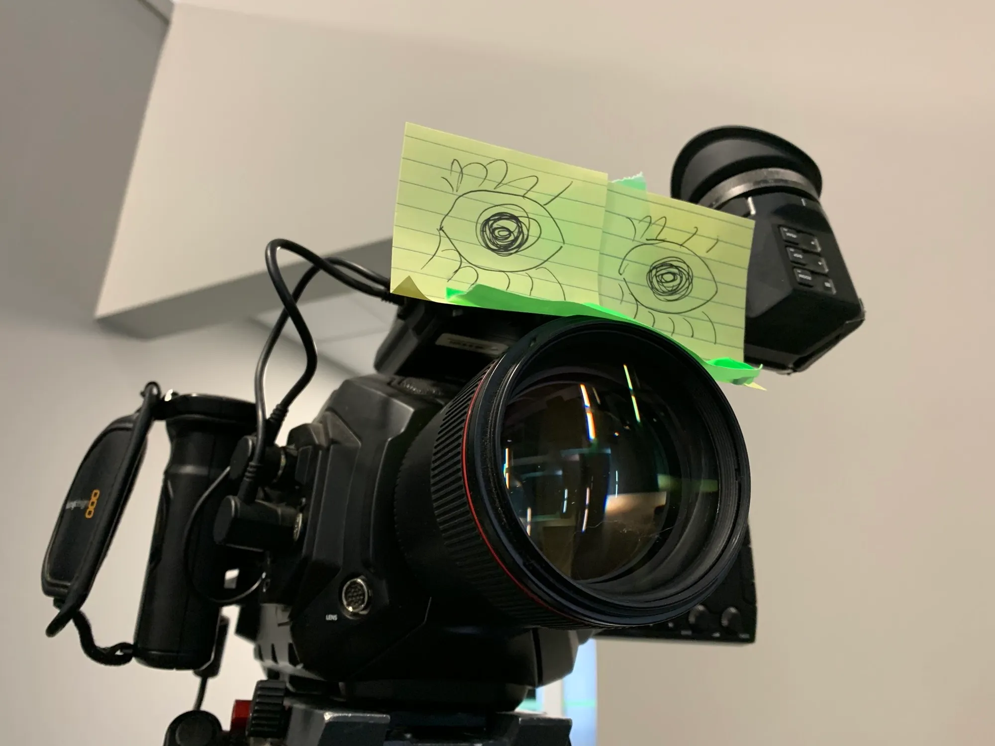 A cinema camera on a tripod with hand-drawn "eyes" sketched on sticky notes taped above the lens.