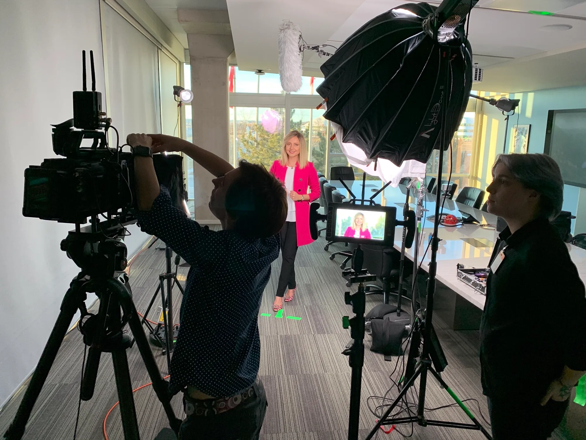 A film crew lighting and shooting a presenter in a pink blazer in a bright office studio.