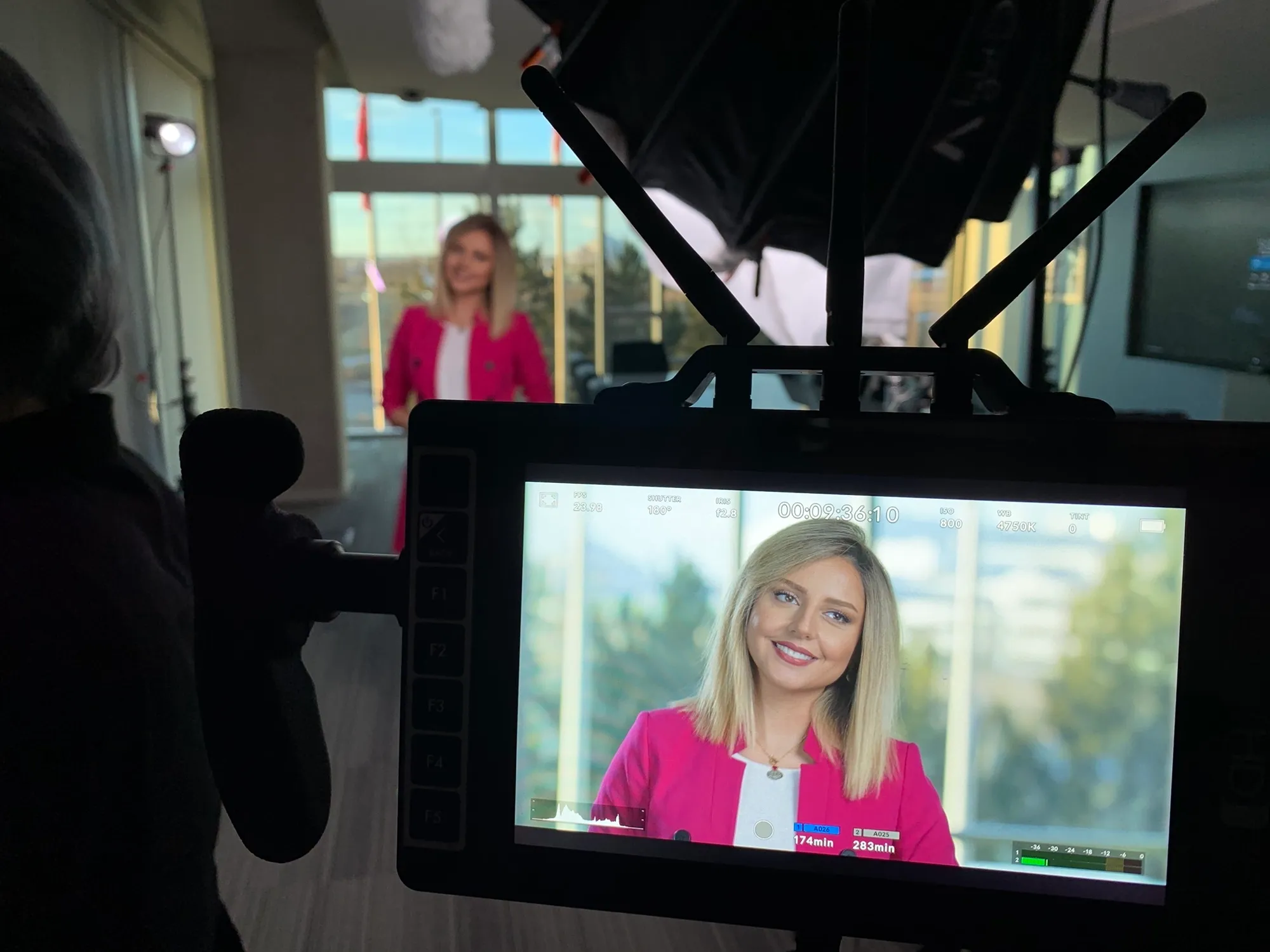 A camera monitor in the foreground showing a close-up of the pink-blazered presenter, with the live set behind it.