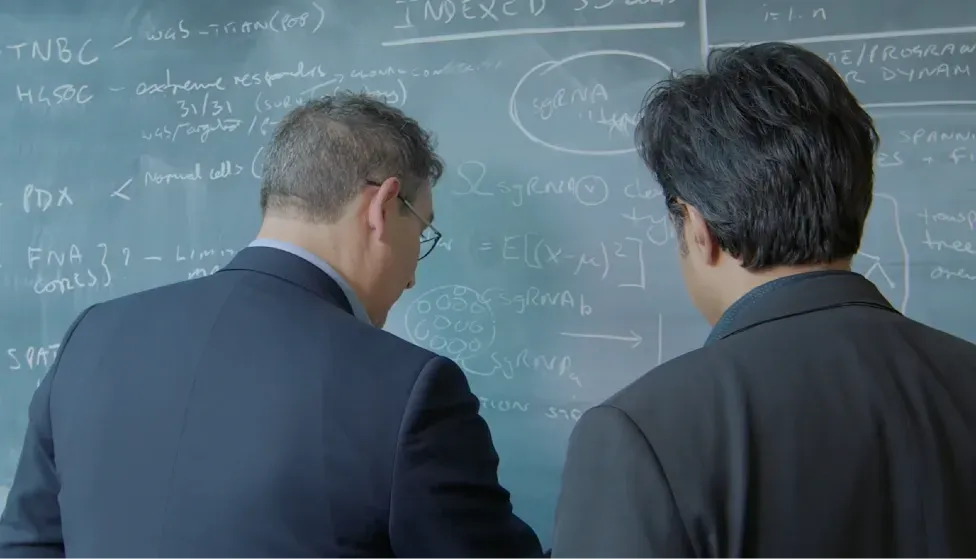 Two researchers study a chalkboard covered in single-cell genomics notation, working through the math behind reading a tumour cell by cell.