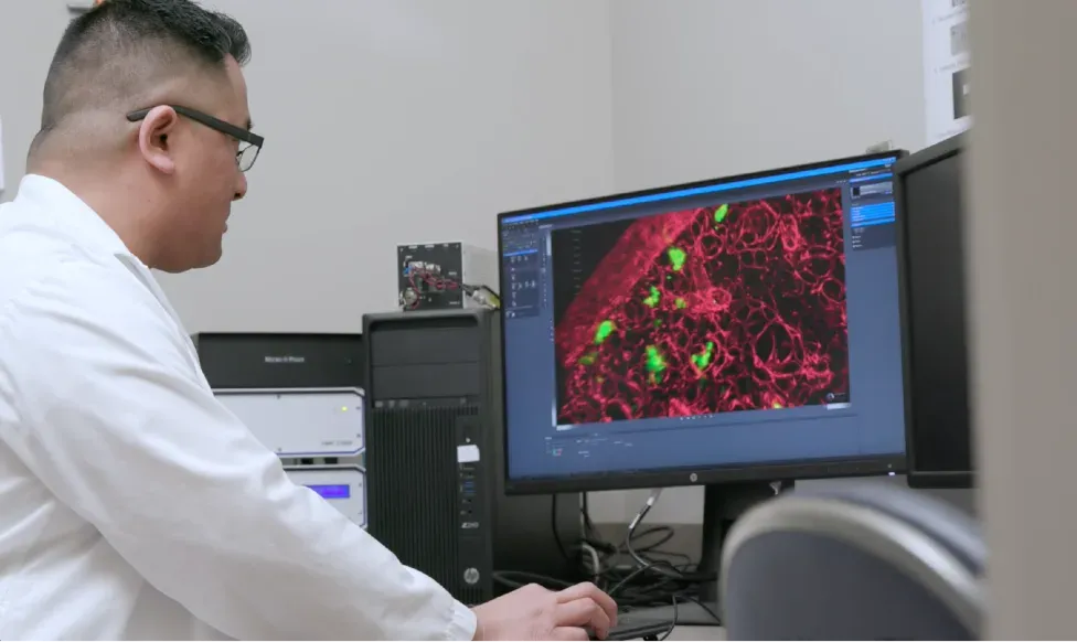 A researcher in a white coat studies fluorescence microscopy on a monitor, cancer tissue rendered in red and green at single-cell resolution.